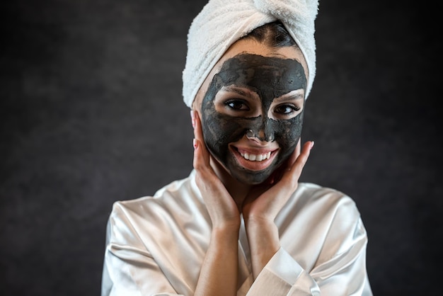 Kaukasische Frau trägt ein weißes Handtuch mit einer nährenden Maske aus schwarzem Tonschlamm im Gesicht, isoliert auf Schwarz