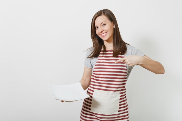 Kaukasische Hausfrau des jungen attraktiven lächelnden Brunette in der gestreiften Schürze, graues T-Shirt lokalisiert. Schöne Haushälterin, die weißen leeren Teller hält