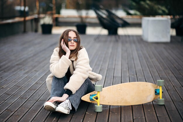 Foto kaukasische hipsterin mit sonnenbrille, pelzmantel und dunklen jeans sitzt auf dem dach neben ihrem skateboard. lieblings-extremhobby einer jungen brünetten