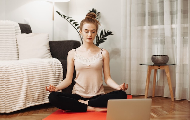 Kaukasische Ingwerdame mit Sommersprossen, die Yoga auf dem Boden unter Verwendung eines Teppichs und eines Laptops tun