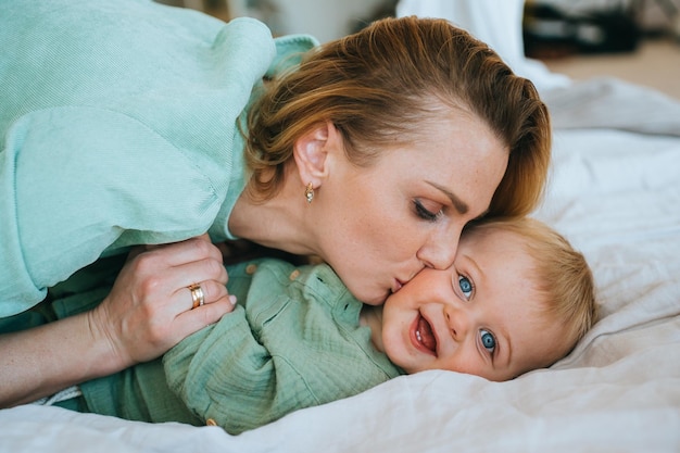 Foto kaukasische junge erwachsene mutter küsst neugeborenen jungen auf die wange. lächelndes baby, das auf dem bett liegt, lächelt