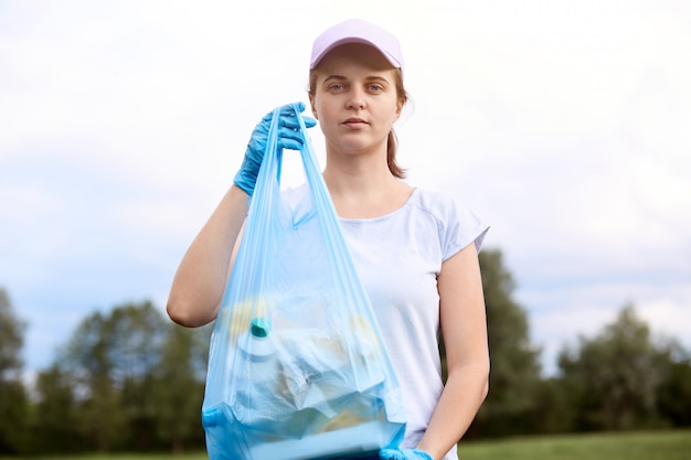 Kaukasische junge Frau, die Müll von der Wiese aufnimmt. Weibliches Reinigungsfeld, Sammeln von Müll im Müllsack, Tragen von T-Shirt und Basiskugelkappe, Stehen mit Bäumen und Himmel i