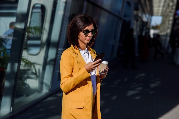 Kaukasische junge Geschäftsfrau, die Smartphone verwendet und Kaffee außerhalb nahe dem Bürozentrum trinkt.