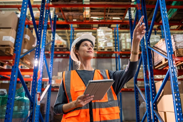 Kaukasische junge Industriearbeiterin, die in einer Produktionsstätte arbeitet, attraktive weibliche Industriefabrikingenieurin, die Helm trägt und Bestellungen und Produkte im Fabriklager glücklich verarbeitet