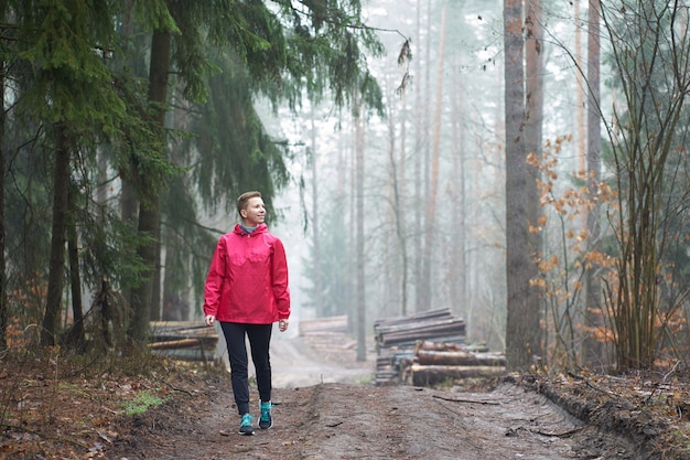 Kaukasische kurzhaarige, getragene lächelnde Frau des rosa Regenmantels, die im nebligen Wald geht