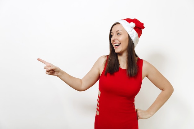 Kaukasische lachende Frau mit charmantem Lächeln in rotem Kleid und Weihnachtsmütze, die wegschaut und mit dem Zeigefinger auf weißem Hintergrund beiseite zeigt. Sankt-Mädchen. Neujahrsfeiertag 2018 Konzept.