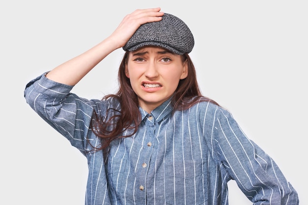 Kaukasische nachdenkliche junge Frau hat einen denkenden Ausdruck, der direkt in die Kamera schaut Zweifelhaftes Mädchen trägt blaues Hemd kann keine Wahl treffen, hat den Ausdruck mit der Hand auf dem Kopf an der weißen Wand in Frage gestellt