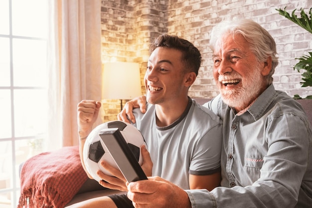 Kaukasische Paare von jungen und älteren Großvater-Fußballfans, die sich ein Fußballspiel im Fernsehen ansehen und auf einem bequemen Sofa im Wohnzimmer sitzen