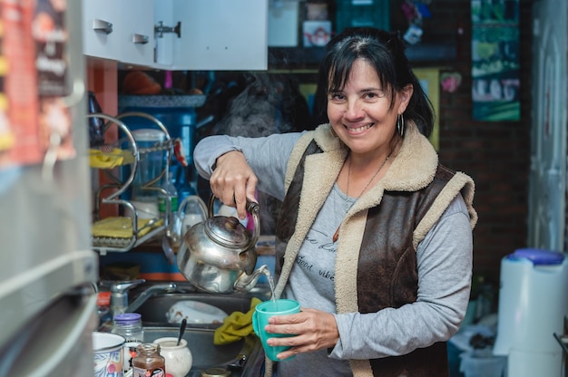 Kaukasische reife Frau, die die Kamera in der Küche anschaut und heißes Wasser in einer Tasse serviert