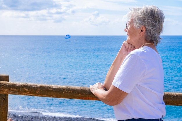 Kaukasische Seniorin, weiß gekleidet, lehnt entspannt an einem Holzbalkon mit Blick auf das Meer und blickt auf den Horizont über Wasser und genießt die Sommerferien