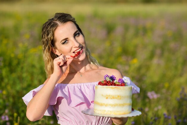 Kaukasische sommersprossige Frau, die einen Kuchen hält und Beeren isst, die an einem sonnigen Tag im Freien posieren.