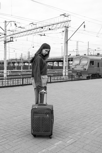 Kaukasische Studentin in heller Kleidung mit einem Koffer, der zum elektrischen Zug am Bahnhof geht, schwarz und weiß. Mädchen mit Koffer am Bahnhof. Eine Reise für Studenten in den Urlaub.