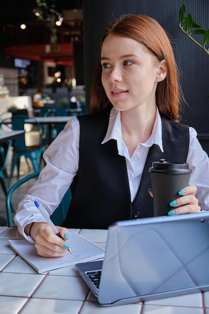 Kaukasische Studentin sitzt am Tisch des Cafés und lernt mit Laptop und macht Hausaufgaben