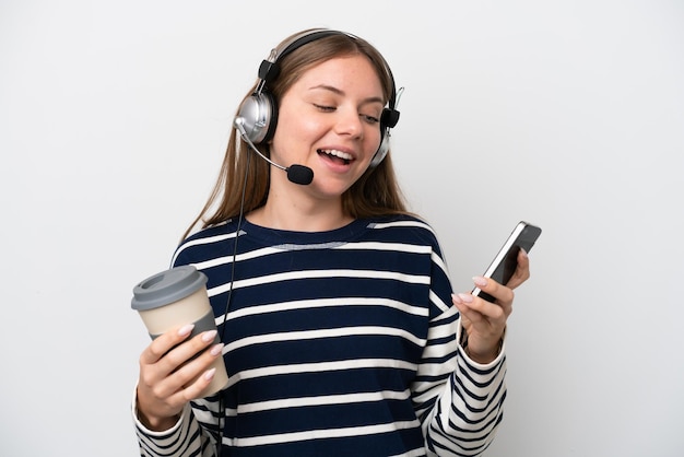 Kaukasische Telemarketerin, die mit einem Headset arbeitet, das auf weißem Hintergrund isoliert ist und Kaffee zum Mitnehmen und ein Handy hält