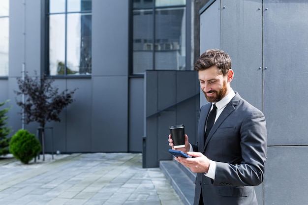 Kaukasischer bärtiger angestellter auf bürogebäude im städtischen hintergrund, der sms-nachrichten schreibt, gute nachrichten handy liest. geschäftsmann, der eine formelle anzugnachricht mit smartphone-handy im freien steht