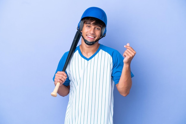 Kaukasischer Baseballspieler mit Helm und Schläger auf blauem Hintergrund isoliert, der Geldgeste macht