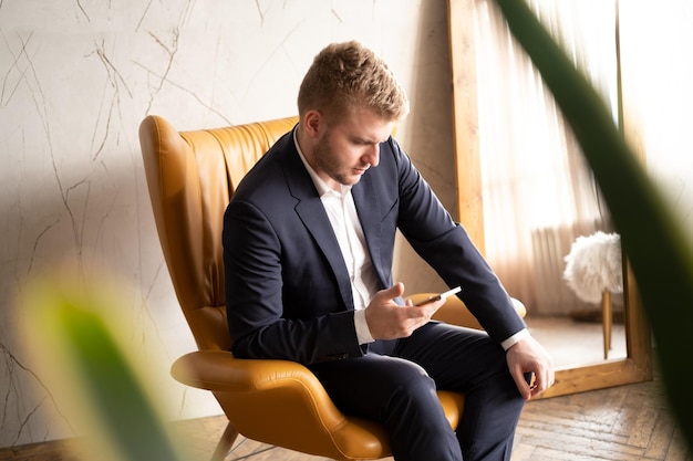 Kaukasischer blonder erwachsener seriöser Mann, der im modernen Interieur mit Smartphone drinnen am Sessel sitztJunger Mann SMS auf dem HandyGeschäftsmann, der das Konzept des Kommunikationsprozesses aushandelt