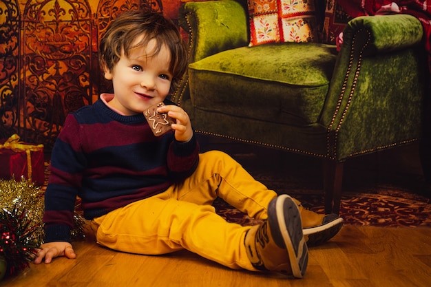 Kaukasischer blonder kleiner Junge mit blauen Augen sitzt und isst einen Keks mit Weihnachtsschmuck