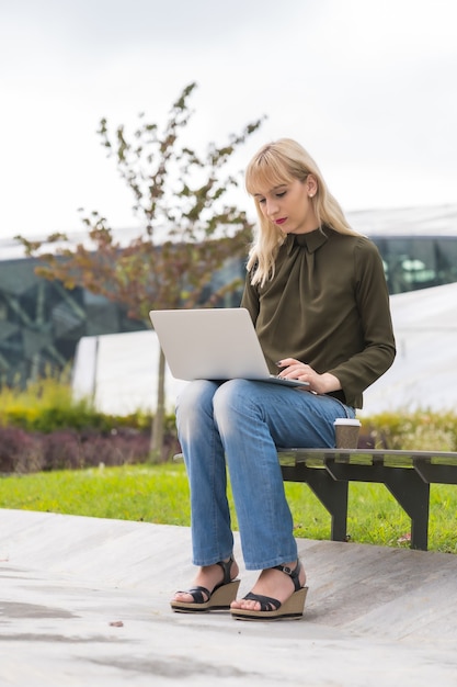 Kaukasischer blonder Mädchenunternehmer-Geschäftspark, der eine E-Mail mit Laptop sendet. Modernes Polygon inmitten der Natur