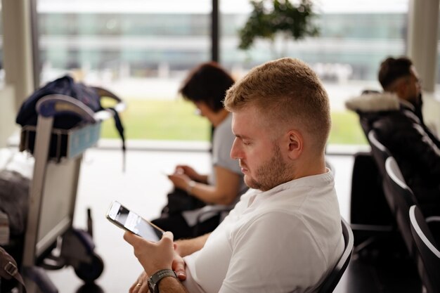 Kaukasischer blonder Mann mittleren Alters, der legere Kleidung trägt und am Flughafen sitzt und auf das Einchecken mit dem Handy wartet