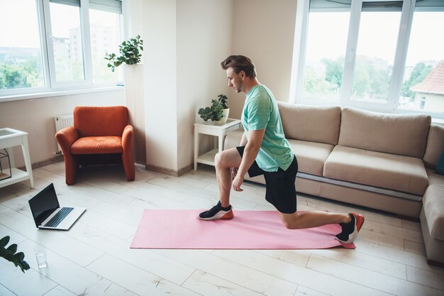 Kaukasischer blonder sportlicher Mann benutzt einen Laptop, während er zu Hause Fitnessübungen macht