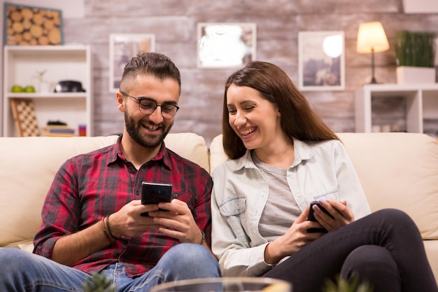 Kaukasischer Freund, der mit seiner Freundin lacht, während er ihr etwas auf seinem Telefon zeigt. Paar sitzt auf der Couch