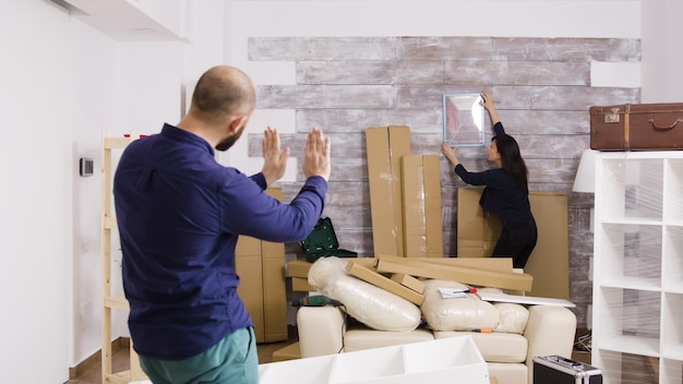 Kaukasischer freund, der seiner freundin sagt, wie man ein bild an der wand ihrer neuen wohnung aufhängt. paare, die hauswand schmücken.