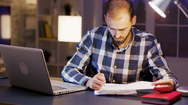 Kaukasischer Geschäftsmann, der ein Hemd trägt, während er Dokumente im Home Office unterschreibt. Laptop auf dem Schreibtisch