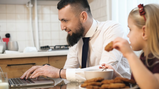 Kaukasischer Geschäftsmann mit Bart, der am Computer arbeitet und seine Tochter, die Kekse am gleichen Tisch isst