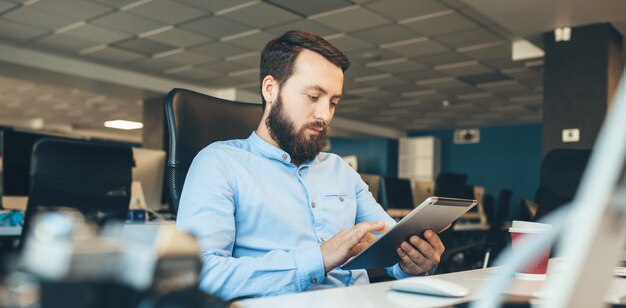 Kaukasischer Geschäftsmann mit Bart, der auf seinem Tablett während der Arbeit im Büro sucht