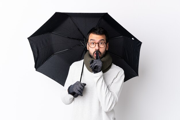 Kaukasischer gutaussehender Mann mit dem Bart, der einen Regenschirm über der lokalisierten weißen Wand zeigt ein Zeichen der Ruhegeste einsetzt Finger in Mund hält