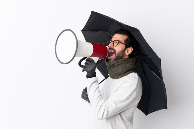 Kaukasischer gutaussehender Mann mit dem Bart, der einen Regenschirm über lokalisierter weißer Wand schreit durch ein Megaphon hält