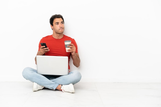 Kaukasischer gutaussehender Mann mit einem Laptop, der auf dem Boden sitzt und Kaffee zum Mitnehmen und ein Handy hält, während er etwas denkt