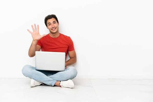 Kaukasischer gutaussehender Mann mit einem Laptop, der auf dem Boden sitzt und mit den Fingern fünf zählt