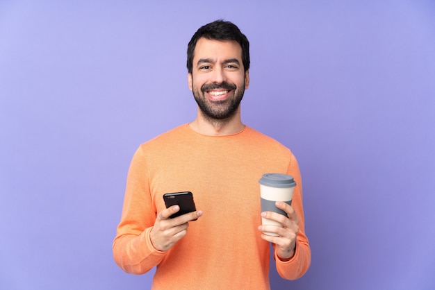 Kaukasischer gutaussehender Mann über isoliertem Purpur, der Kaffee hält, zum wegnehmen und ein Handy