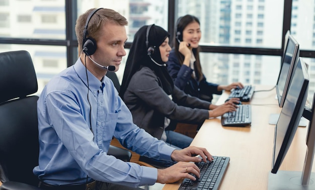 Kaukasischer gutaussehender Mann und eine Gruppe von verschiedenen Telemarketing-Kundendienstmitarbeitern im Callcenter. Call-Center-Mitarbeiter, begleitet von Team. Lächeln des Kundendienstmitarbeiters bei der Arbeit.