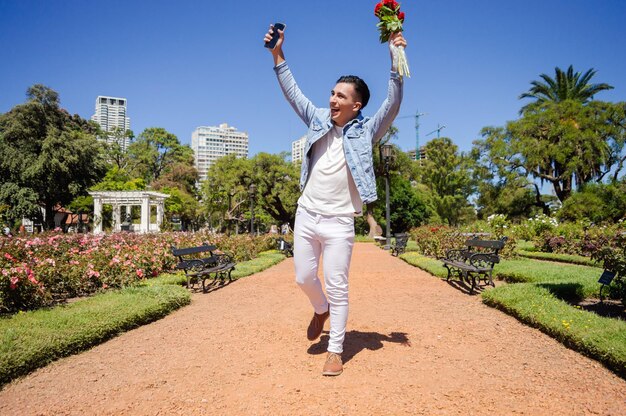 Kaukasischer hispanischer lateinamerikanischer argentinischer Mann, der in einen Blumenstrauß in der Hand verliebt ist, sehr glücklich, das Springen auf einem Fuß in einem Quadrat zu feiern, verärgert lächelnd mit guten Nachrichten