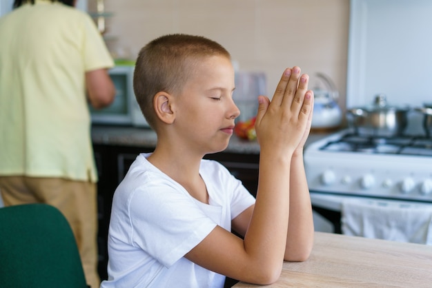 Kaukasischer Junge betet zu Hause am Tisch, bevor er isst, bleib zu Hause und bete zu Gott Online-Kirchenanbetung o ...