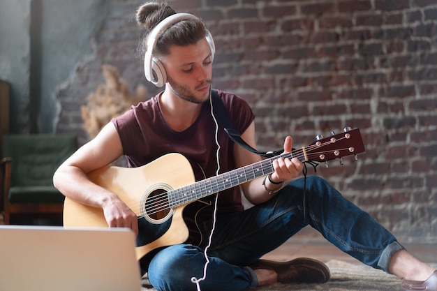 Kaukasischer junger Erwachsener lernt Gitarre Online-Musikunterricht mit entferntem Lehrer-Computer-Mobilgerät Gutaussehender Hipster-Mann, der Musik durch Laptop-Fernunterricht-Lernkonzept unterrichtet