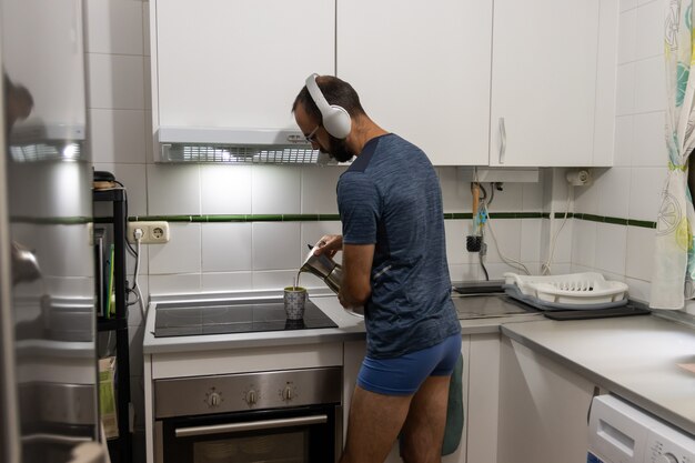 Kaukasischer junger Mann, der zu Hause Kaffee in der Küche serviert, während er mit Kopfhörern Musik hört