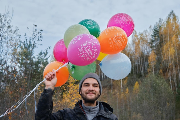 Kaukasischer junger Mann hält viele Heliumballons draußen und lächelt Feiern Sie Geburtstag in der Natur