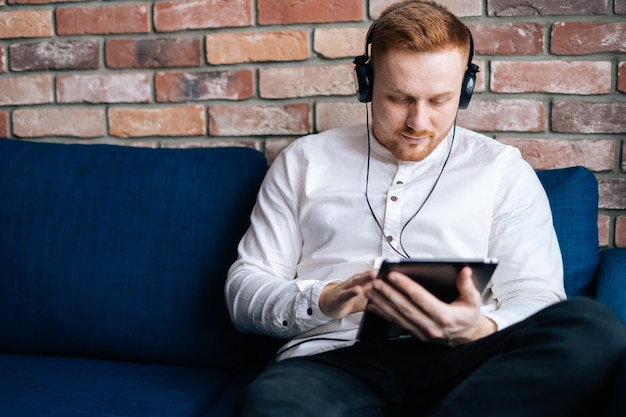 Kaukasischer junger Mann mit Kopfhörern, der einen Film auf einem Tablet-Computer ansieht, während er zu Hause auf der Couch sitzt. Konzept der Entspannung zu Hause.