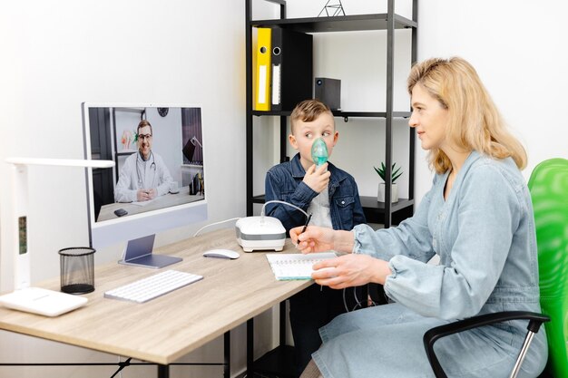 Kaukasischer kleiner Junge, der zu Hause mit einem Vernebler inhaliert Eine junge Mutter hilft ihrem Sohn bei einem Online-Meeting mit einem Arzt, mit einem Vernebler zu inhalieren