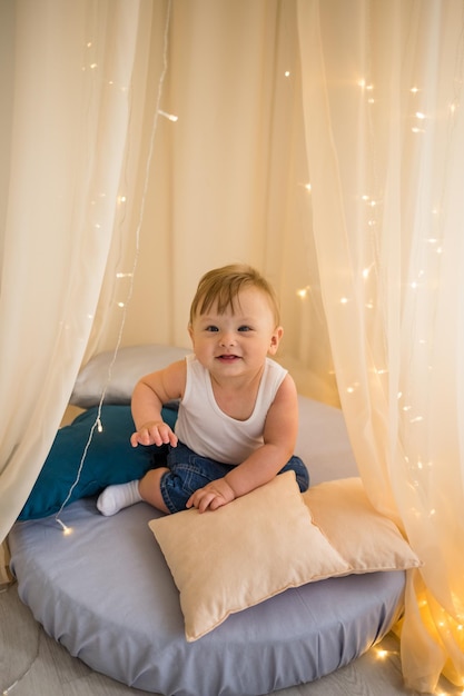 Foto kaukasischer kleiner junge sitzt auf einer matratze unter einem baldachin und spielt mit kissen ein verspielter kleiner junge in einem weißen body und jeans sitzt im spielzimmer