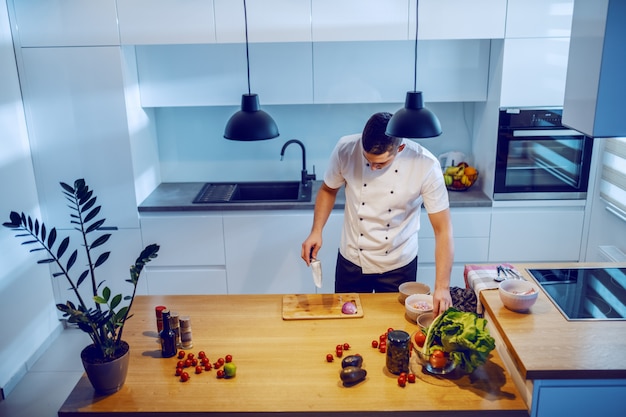 kaukasischer Koch in der Uniform, die in der Küche steht, Messer hält und Abendessen vorbereitet