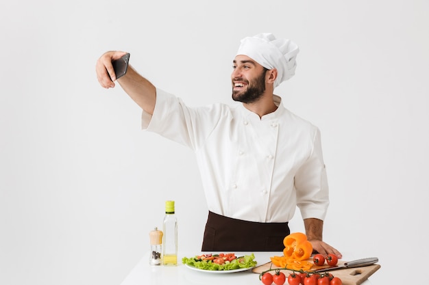 Kaukasischer Kochmann in Uniform, der bei der Arbeit ein Selfie-Foto von Gemüsesalat auf dem Smartphone macht, isoliert über weißer Wand?