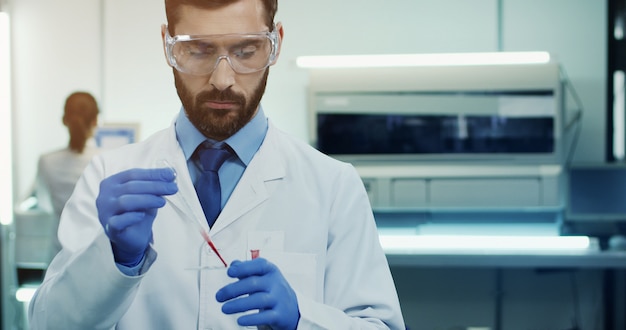 Kaukasischer Laborwissenschaftler des jungen Mannes in den Gläsern, die eine Blutprobe mit einem Gefäß in den Händen bilden. Porträt. Nahansicht