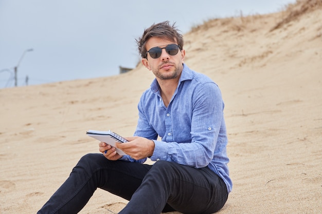 Kaukasischer Mann, der auf dem Sand am Strand sitzt, eine Brille trägt und ein Notizbuch hält, in das er Gedichte schreiben kann.