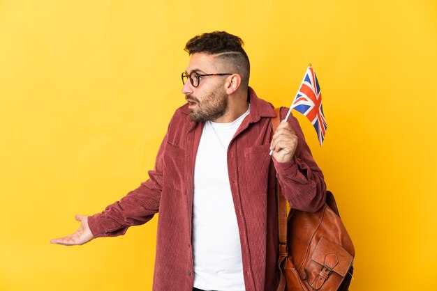 Kaukasischer Mann, der eine Flagge des Vereinigten Königreichs hält, die auf gelbem Hintergrund mit Überraschungsausdruck isoliert ist, während er zur Seite schaut