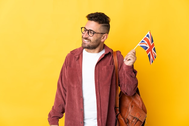Kaukasischer Mann, der eine Flagge des Vereinigten Königreichs lokalisiert auf gelbem Blick zur Seite und lächelnd hält